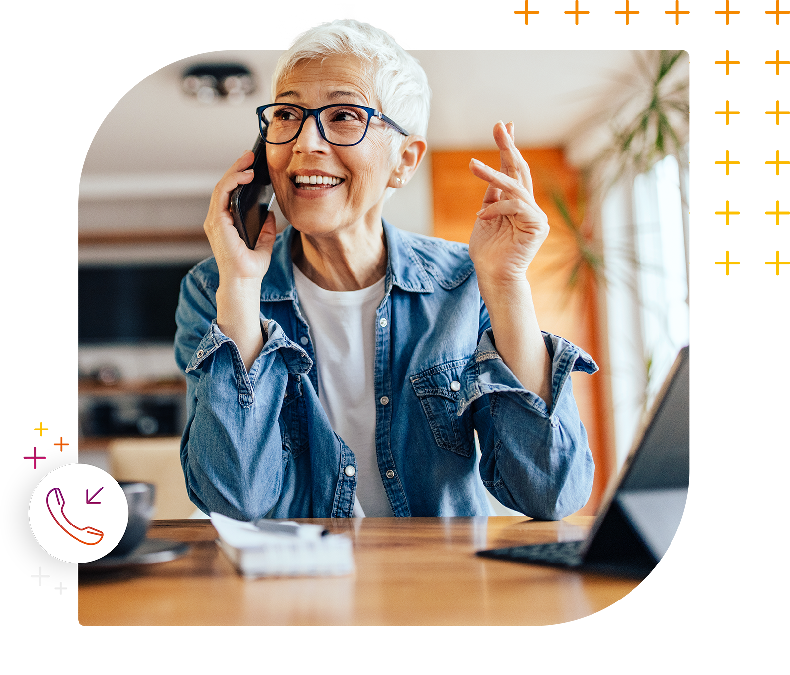 Ältere Frau mit kurzen weißen Haaren und Brille telefoniert lächelnd, während sie an einem Holztisch sitzt. Ein Laptop und Notizblock liegen vor ihr.