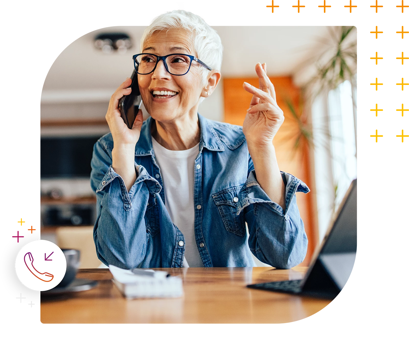 Ältere Frau mit kurzen weißen Haaren und Brille telefoniert lächelnd, während sie an einem Holztisch sitzt. Ein Laptop und Notizblock liegen vor ihr.